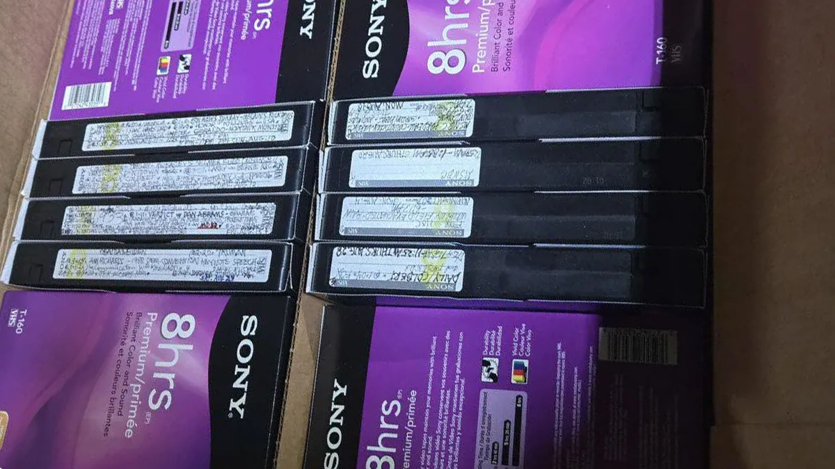 نمایی از نوار کاستهای VHS سونی با بستهبندی بنفش که در داخل یک جعبه چیده شدهاند و برچسبهای دستنویس روی کناره کاستها به سختی قابل مشاهده است.