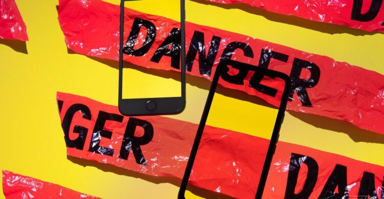 دو گوشی هوشمند مشکی رنگ که به صورت مورب روی پسزمینه زرد قرار گرفتهاند و نوار هشدار پلاستیکی قرمز رنگ با کلمه "DANGER" دور آنها پیچیده شده است.