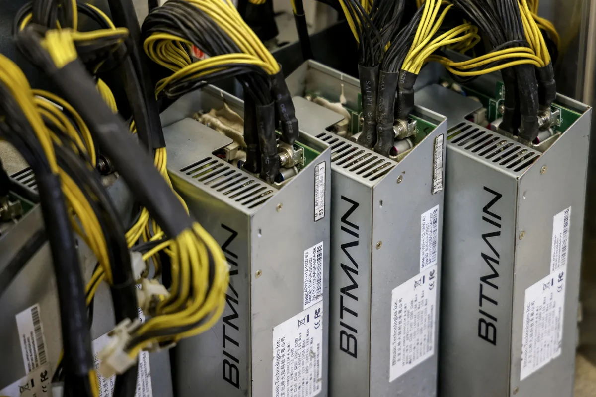 نمای نزدیک از بخش‌های کناری دستگاه‌های ماینینگ بیت‌کوین Bitmain که نام "BITMAIN" روی آنها حک شده و تعداد زیادی کابل زرد و سیاه از بالا به آنها متصل شده است.