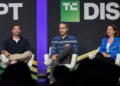 سه سخنران روی صندلیها در یک پنل گفتگو در کنفرانس TechCrunch Disrupt، که در پسزمینه یک بنر با لوگوی "DISRUPT" دیده میشود.