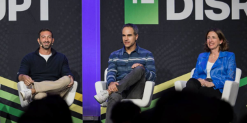 سه سخنران روی صندلی‌ها در یک پنل گفتگو در کنفرانس TechCrunch Disrupt، که در پس‌زمینه یک بنر با لوگوی "DISRUPT" دیده می‌شود.