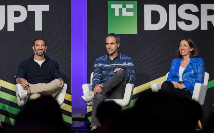سه سخنران روی صندلیها در یک پنل گفتگو در کنفرانس TechCrunch Disrupt، که در پسزمینه یک بنر با لوگوی "DISRUPT" دیده میشود.