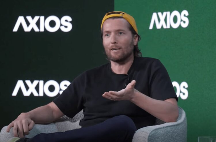 الکس کارپ (Alex Karp)، مدیرعامل پالتیر (Palantir)، با کلاه زرد و تی&zwnj;شرت مشکی روی صندلی در حال صحبت کردن در یک رویداد Axios.