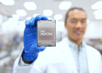 یک مرد با روپوش سفید و دستکش آبی، تراشه Intel Xeon را که Heat Spreader آن رو به دوربین است، در یک آزمایشگاه نگه داشته است.