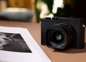 دوربین Leica Q3 Monochrom به رنگ سیاه، که با یک هود لنز روی یک سطح قهوه‌ای در کنار مجموعه‌ای از عکس‌های چاپ شده سیاه و سفید قرار گرفته است.