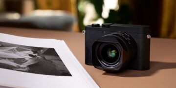 دوربین Leica Q3 Monochrom به رنگ سیاه، که با یک هود لنز روی یک سطح قهوه‌ای در کنار مجموعه‌ای از عکس‌های چاپ شده سیاه و سفید قرار گرفته است.