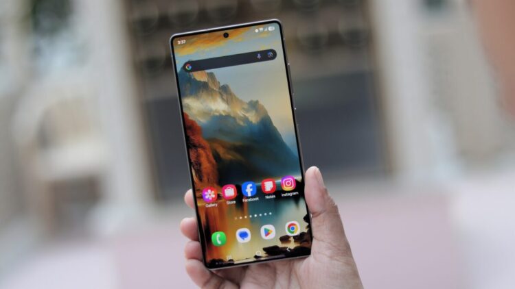 یک دست گوشی هوشمند سامسونگ با صفحه‌نمایش روشن و والپیپر کوهستانی را نگه داشته است.