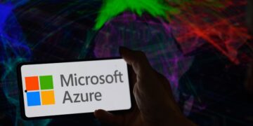 لوگوی Microsoft Azure روی صفحه یک گوشی هوشمند که توسط دست در مقابل یک پس‌زمینه رنگارنگ و انتزاعی از نور نگه داشته شده است.