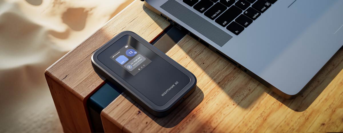 یک هاتاسپات (Hotspot) موبایل Netgear Nighthawk 5G در کنار یک لپتاپ روی یک میز چوبی در یک محیط روشن با شن در پسزمینه.