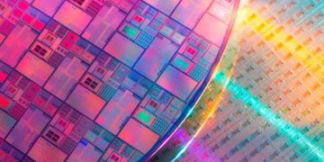 نمای نزدیک از یک ویفر سیلیکونی بزرگ با طراحی‌های مدار میکروسکوپی در رنگ‌های متالیک روشن صورتی، آبی و نارنجی.