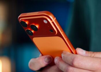 رندر گوشی iPhone 18 Pro نارنجی با ماژول دوربین سهگانه و فریم نارنجی براق که در دست یک فرد با نورپردازی هنری نگه داشته شده است.