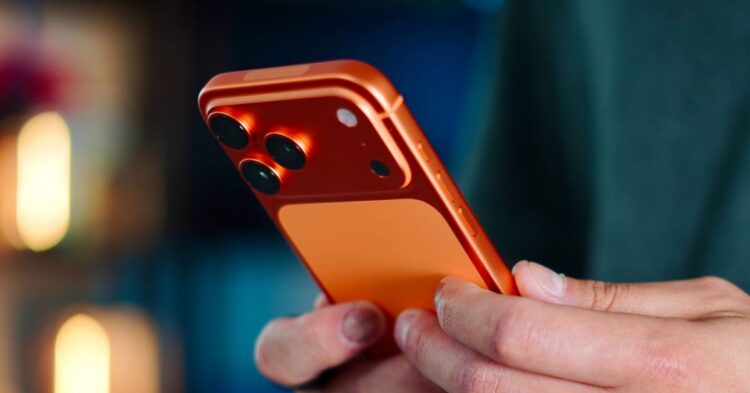 رندر گوشی iPhone 18 Pro نارنجی با ماژول دوربین سه‌گانه و فریم نارنجی براق که در دست یک فرد با نورپردازی هنری نگه داشته شده است.