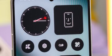 نمای نزدیک از یک گوشی هوشمند (احتمالاً Nothing Phone) که ویجت‌های ساعت آنالوگ و طرحی پیکسلی را در یک محیط تیره و مینیمال نشان می‌دهد.