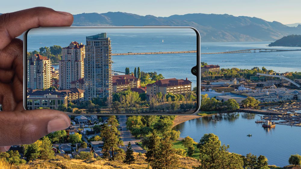 یک دست که یک گوشی هوشمند را نگه داشته و نمای نزدیک از یک شهر ساحلی (شامل ساختمان‌های بلند و دریاچه) را روی صفحه نمایش می‌دهد، در حالی که پس‌زمینه خود همان منظره را در حالت وسیع‌تر نشان می‌دهد.
