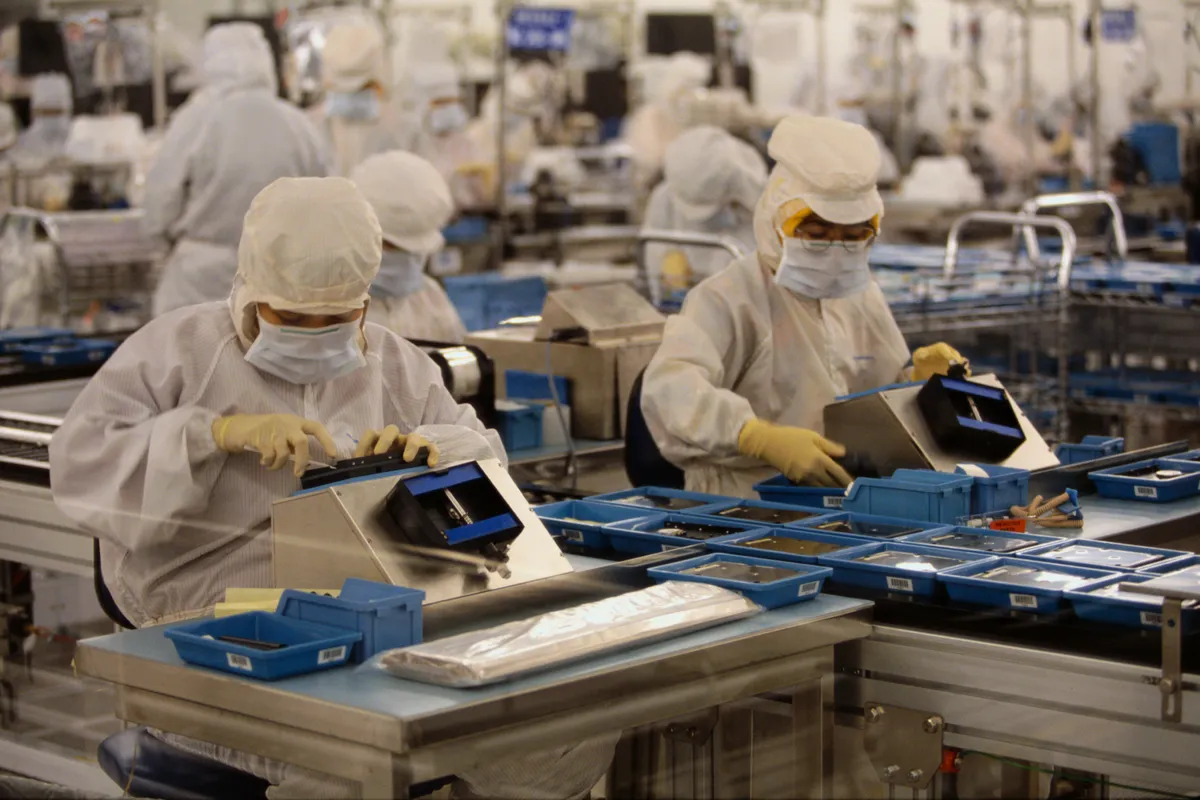 کارگران در اتاق تمیز یک کارخانه تولید نیمه‌هادی یا حافظه، که لباس‌های محافظ سفید و ماسک بهداشتی پوشیده‌اند و در حال مونتاژ قطعات بر روی میز کار هستند.