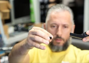 یک مرد ریشو که قطعه شکسته یک کانکتور کارت گرافیک را در یک دست و بخش اصلی کانکتور را در دست دیگر گرفته است.