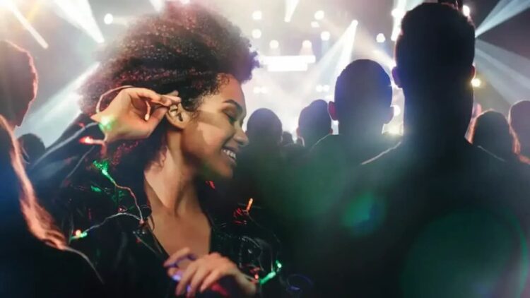 یک زن با موهای فر و لباس براق در حال لبخند زدن و لذت بردن از یک کنسرت یا رویداد موسیقی شلوغ، که دست خود را به گوشش نزدیک کرده است.