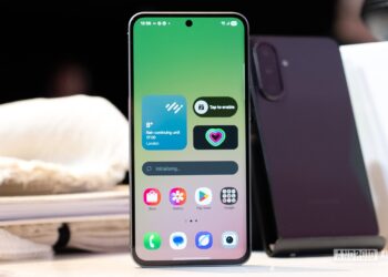 یک گوشی هوشمند (احتمالاً نمونه اولیه Galaxy S26) که روی میز قرار دارد و صفحه اصلی آن ویجت‌های آب و هوا و ضربان قلب را با پس‌زمینه گرادیانتی سبز و رابط کاربری One UI نشان می‌دهد.