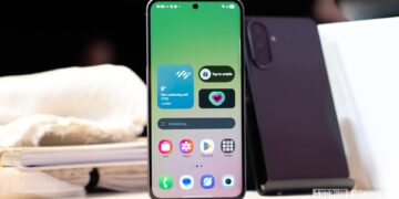 یک گوشی هوشمند (احتمالاً نمونه اولیه Galaxy S26) که روی میز قرار دارد و صفحه اصلی آن ویجتهای آب و هوا و ضربان قلب را با پسزمینه گرادیانتی سبز و رابط کاربری One UI نشان میدهد.