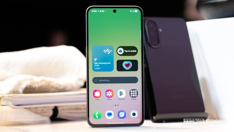 یک گوشی هوشمند (احتمالاً نمونه اولیه Galaxy S26) که روی میز قرار دارد و صفحه اصلی آن ویجتهای آب و هوا و ضربان قلب را با پسزمینه گرادیانتی سبز و رابط کاربری One UI نشان میدهد.