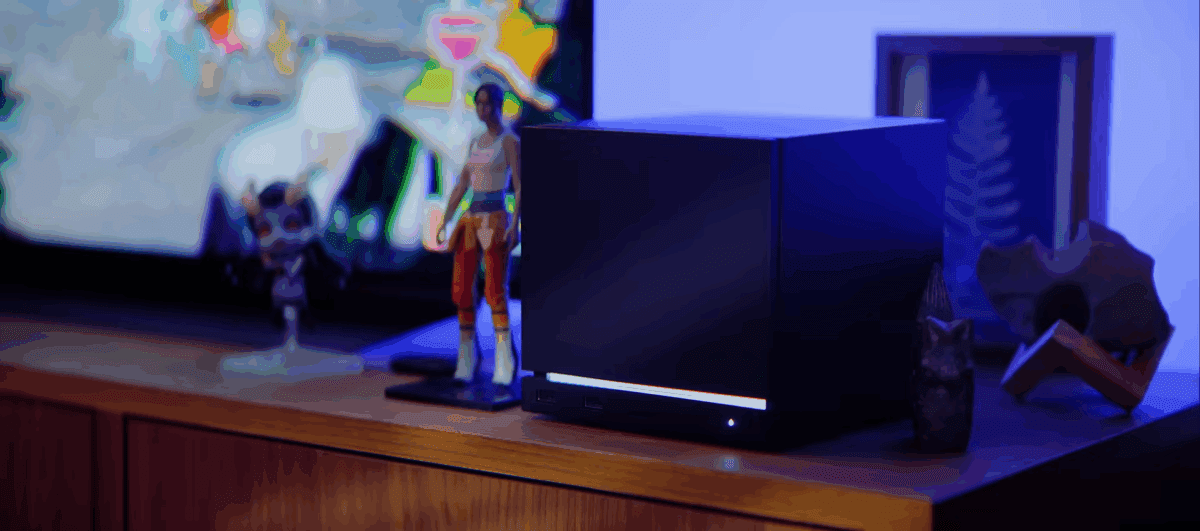 یک سیستم Steam Machine مکعبی و مشکی رنگ روی یک میز چوبی، که یک تلویزیون و مجسمههای شخصیتی بازیهای ویدیویی در پسزمینه آن دیده میشوند.