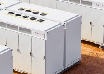 ردیف‌هایی از واحدهای بزرگ ذخیره‌سازی باتری Tesla Megapack سفید رنگ با فن‌های خنک‌کننده در بالا، که در یک سایت خاکی در فضای باز قرار دارند.