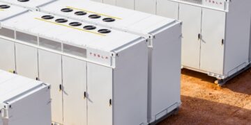 ردیفهایی از واحدهای بزرگ ذخیرهسازی باتری Tesla Megapack سفید رنگ با فنهای خنککننده در بالا، که در یک سایت خاکی در فضای باز قرار دارند.