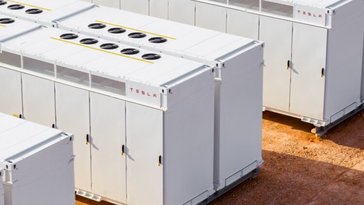 ردیف‌هایی از واحدهای بزرگ ذخیره‌سازی باتری Tesla Megapack سفید رنگ با فن‌های خنک‌کننده در بالا، که در یک سایت خاکی در فضای باز قرار دارند.