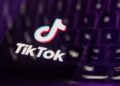 لوگوی TikTok و متن "TikTok" روی صفحه نمایش گوشی یا لپ‌تاپ که در محیطی با نورپردازی بنفش و تاریک به صورت فوکوس نشده قرار دارد.