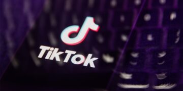 لوگوی TikTok و متن "TikTok" روی صفحه نمایش گوشی یا لپ‌تاپ که در محیطی با نورپردازی بنفش و تاریک به صورت فوکوس نشده قرار دارد.