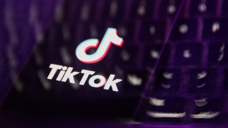 لوگوی TikTok و متن "TikTok" روی صفحه نمایش گوشی یا لپتاپ که در محیطی با نورپردازی بنفش و تاریک به صورت فوکوس نشده قرار دارد.