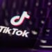 لوگوی TikTok و متن "TikTok" روی صفحه نمایش گوشی یا لپ‌تاپ که در محیطی با نورپردازی بنفش و تاریک به صورت فوکوس نشده قرار دارد.