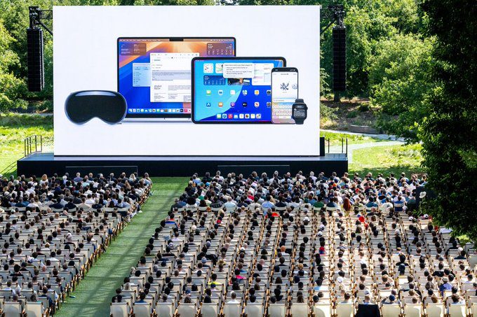 نمای کلی از یک رویداد اپل در فضای باز، که جمعیتی بزرگ روی صندلی‌ها نشسته‌اند و یک صفحه نمایش بزرگ محصولات (هدست Vision Pro، مک‌بوک، آیپد و آیفون) را نشان می‌دهد.