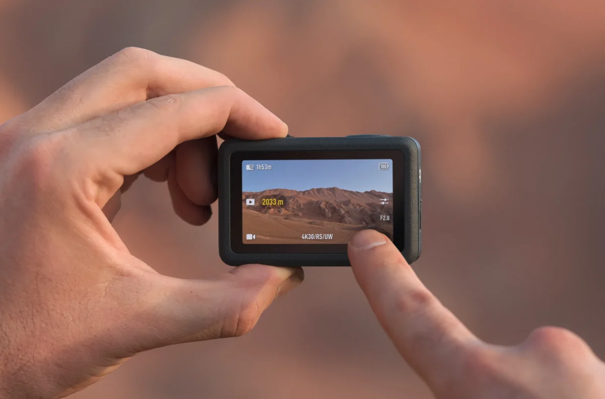یک دست که دوربین اکشن DJI را نگه داشته و با انگشت روی صفحه نمایش لمس می‌کند؛ صفحه نمایش منظره کوهستانی و تنظیمات فیلم‌برداری را نمایش می‌دهد.