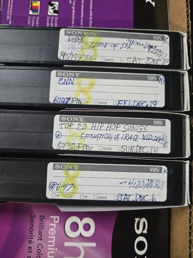 ردیفی از نوار کاستهای VHS سونی که برچسبهای دستنویس با محتویاتی مانند "FOX SOME OF SNL"، "CNN" و "TOP 20 HIP HOP SONGS" روی آنها دیده میشود.