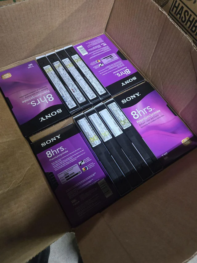 درون یک جعبه مقوایی باز که پر از نوار کاستهای VHS سونی با قابهای بنفش رنگ ۸ ساعته است.