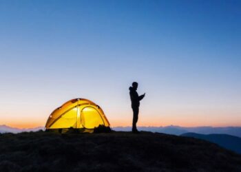 تصویری از یک فرد که کنار چادر کمپاش در محلی بدون تکنولوژی دارد از گوشی استفاده میکند