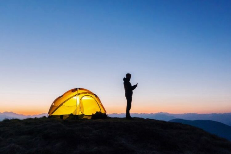 تصویری از یک فرد که کنار چادر کمپ‌اش در محلی بدون تکنولوژی دارد از گوشی استفاده می‌کند