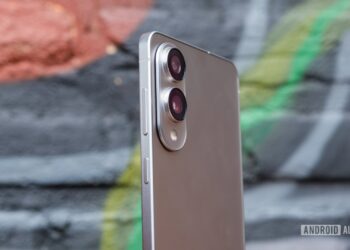 نمای جانبی گوشی هوشمند با بدنه نقرهای و ماژول دوربین دوگانه بزرگ و دکمههای کناری، که در مقابل یک دیوار گرافیتی قرار گرفته است.