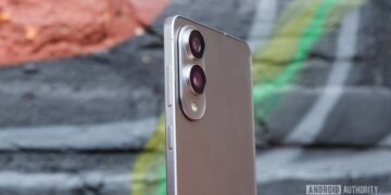 نمای جانبی گوشی هوشمند با بدنه نقرهای و ماژول دوربین دوگانه بزرگ و دکمههای کناری، که در مقابل یک دیوار گرافیتی قرار گرفته است.