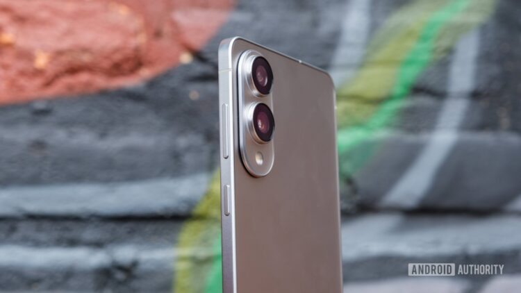 نمای جانبی گوشی هوشمند با بدنه نقرهای و ماژول دوربین دوگانه بزرگ و دکمههای کناری، که در مقابل یک دیوار گرافیتی قرار گرفته است.