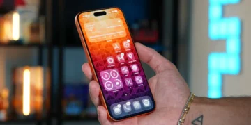 یک دست در حال نگه داشتن یک گوشی هوشمند نارنجی با صفحه نمایش روشن که یک والپیپر هندسی رنگارنگ و آیکون‌های برنامه را نشان می‌دهد.