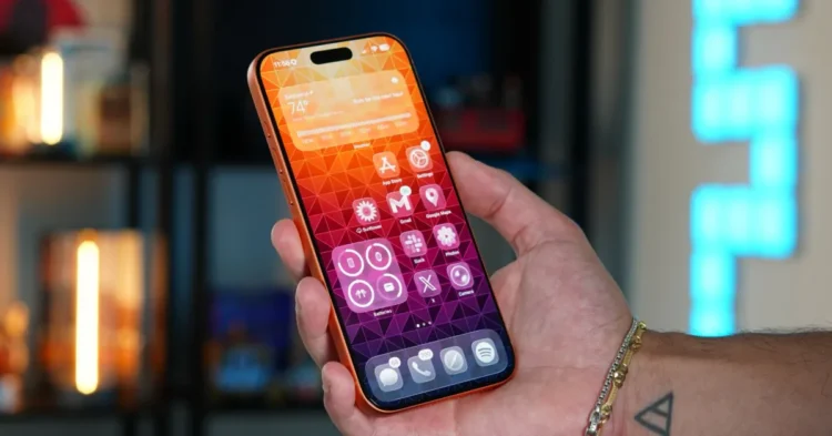 یک دست در حال نگه داشتن یک گوشی هوشمند نارنجی با صفحه نمایش روشن که یک والپیپر هندسی رنگارنگ و آیکونهای برنامه را نشان میدهد.