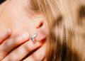 نمای نزدیک از یک زن با موهای بلوند که یک حلقه نقرهای یا حسگر پوشیدنی در لاله گوشش دارد و با دست روی صورت خود را پوشانده است.