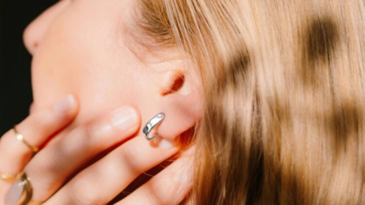 نمای نزدیک از یک زن با موهای بلوند که یک حلقه نقره‌ای یا حسگر پوشیدنی در لاله گوشش دارد و با دست روی صورت خود را پوشانده است.