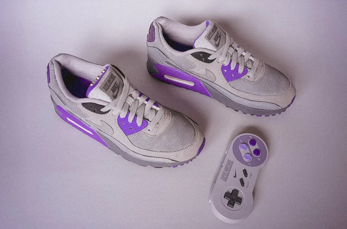 یک کفش ورزشی Nike Air Max 90 با تم Super Nintendo Entertainment System (SNES) به رنگهای خاکستری و بنفش، که یک کنترلر SNES در کنار آن قرار دارد.