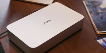 مینی پی‌سی Olares One به رنگ سفید/نقره‌ای با لوگوی "Olares" در بالا، که روی یک میز چوبی در کنار یک لپ‌تاپ و کتاب قرار گرفته است.