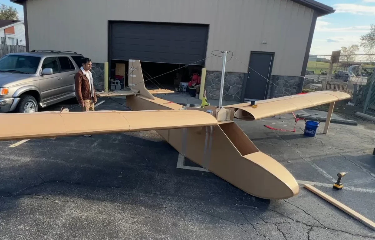 نمای بالایی از هواپیمای مقوایی دست‌ساز در محوطه‌ای باز کنار یک سوله، در حالی که بال‌ها و بدنهٔ مقوایی در مرحلهٔ آماده‌سازی نهایی قرار دارند.