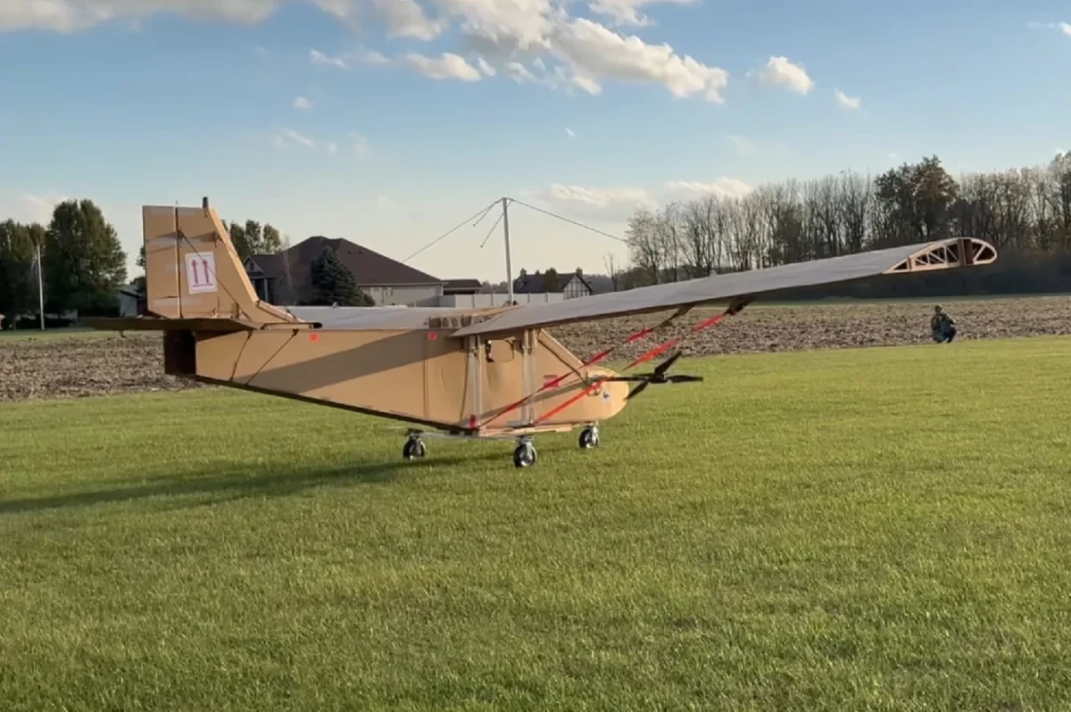 هواپیمای دست‌ساز مقوایی در ابعاد انسانی روی زمین چمن‌زار قرار دارد و سازهٔ بدنه و بال‌های آن پیش از پرواز آزمایشی دیده می‌شود.