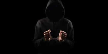 نمای سیلوئت یک فرد با هودی تیره که دستبند فلزی به دستان دارد، در پسزمینه کاملاً مشکی، تصویر مفهومی از جرم سایبری، بازداشت یا فعالیت غیرقانونی دیجیتال